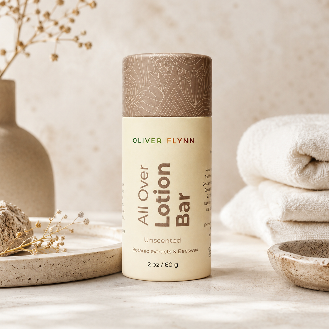 Oliver Flynn All Over Lotion Bar in a beige and brown cylindrical container, styled on a light stone surface with a folded towel, ceramic bowl, and dried flowers in the background.
